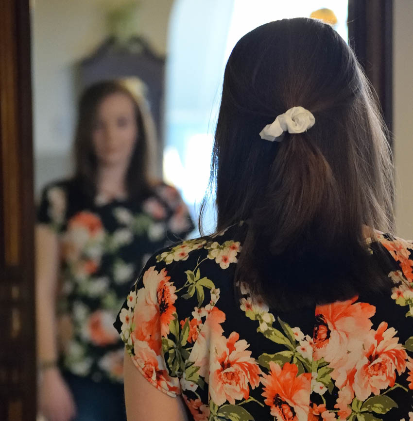 Woman in a flower dress, viewed from behind, looking at an out-of-focus image of herself in a mirror.