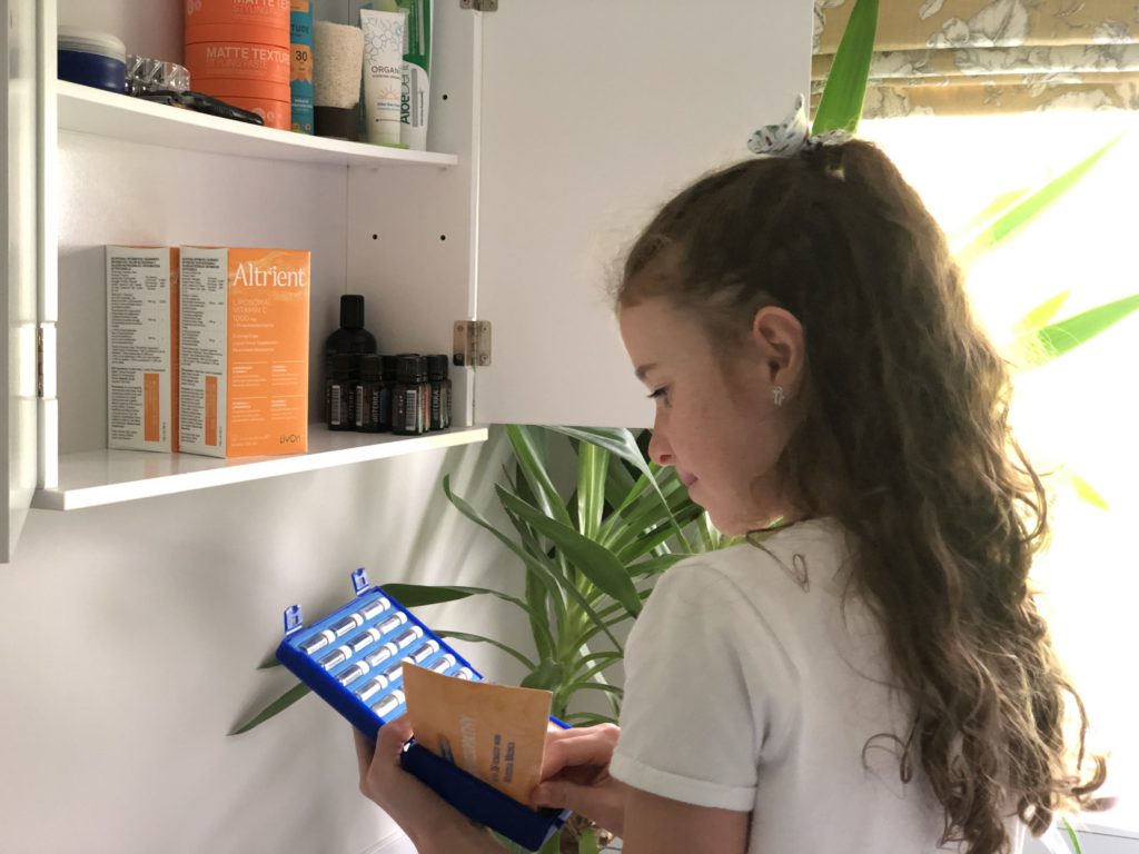 A little girl choosing a Helios homeopathy remedy with a medicine cabinet in the background which includes Altrient vitamin C and DoTerra essential oils